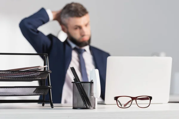 Очки и канцелярские принадлежности на столе рядом задумчивый бизнесмен и ноутбук на размытом фоне — стоковое фото