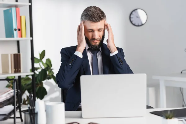 Empresario que sufre de dolor de cabeza cerca de la computadora portátil en primer plano borroso en la oficina - foto de stock