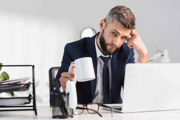 Uomo d'affari con tazza vicino a laptop, occhiali e cancelleria in primo piano sfocato in ufficio — Foto stock