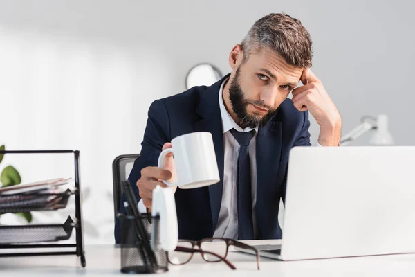 Businessman holding cup near laptop, eyeglasses and stationery on blurred foreground in office — Stock Photo