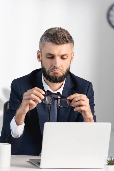 Sad businessman holding eyeglasses near laptop in office — Stock Photo