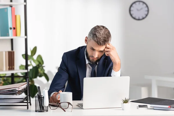 Empresário cansado olhando para laptop perto de papelaria, xícara e óculos em primeiro plano desfocado — Fotografia de Stock