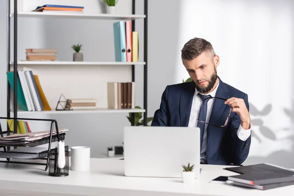 Businessman holding eyeglasses near laptop and papers on blurred foreground in office — Stock Photo