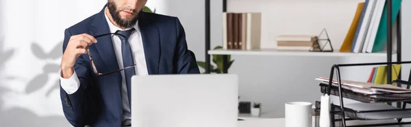 Vue recadrée de l'homme d'affaires tenant des lunettes près d'un ordinateur portable sur le premier plan flou, bannière — Photo de stock