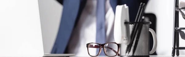 Eyeglasses near stationery and laptop on office table with businessman on blurred background, banner — Stock Photo