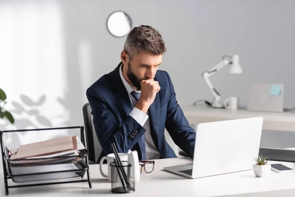 Konzentrierter Geschäftsmann blickt im Büro auf Laptop in der Nähe von Dokumenten und Schreibwaren im verschwommenen Vordergrund — Stockfoto