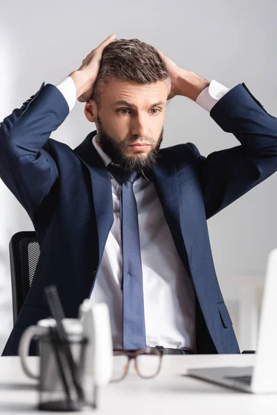 Homme d'affaires fatigué avec les mains près de la tête regardant ordinateur portable sur le premier plan flou au bureau — Photo de stock