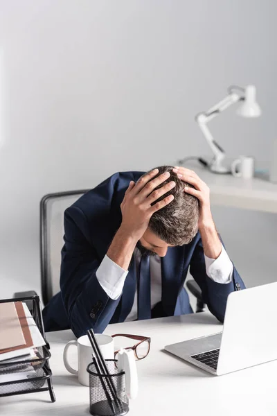 Empresário cansado com as mãos perto da cabeça sentado ao lado do laptop e artigos de papelaria na mesa no escritório — Fotografia de Stock