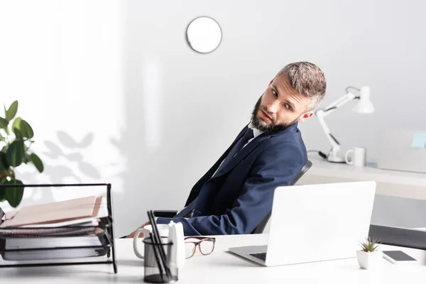 Homme d'affaires regardant ordinateur portable près des lunettes et des documents sur le premier plan flou dans le bureau — Photo de stock