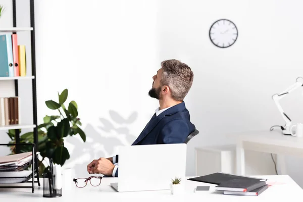 Бизнесмен смотрит в сторону, сидя рядом с гаджетами и ноутбуками на размытом переднем плане в офисе — стоковое фото