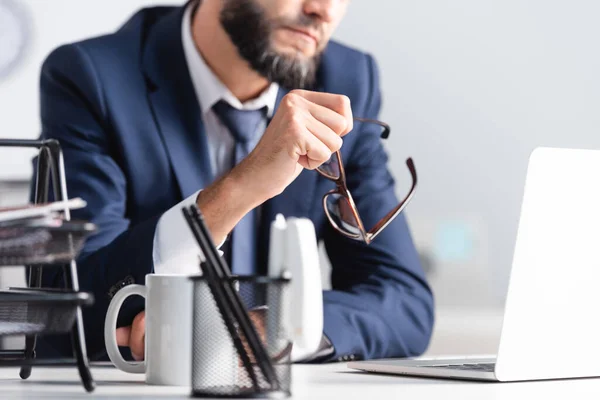 Vista recortada del hombre de negocios sosteniendo anteojos cerca de la computadora portátil y la taza en primer plano borrosa - foto de stock