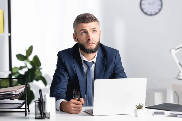 Pensive businessman holding eyeglasses while looking away near laptop and papers on table — Stock Photo