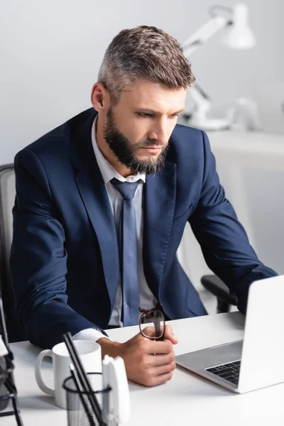 Homem de negócios barbudo segurando óculos ao usar laptop perto da xícara na mesa de trabalho — Fotografia de Stock