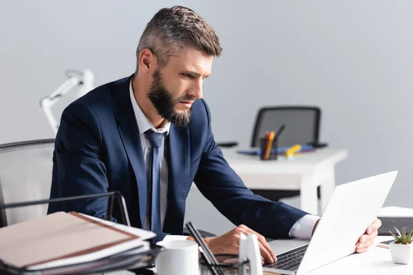 Empresário usando laptop perto de documentos e xícara em primeiro plano desfocado no escritório — Fotografia de Stock