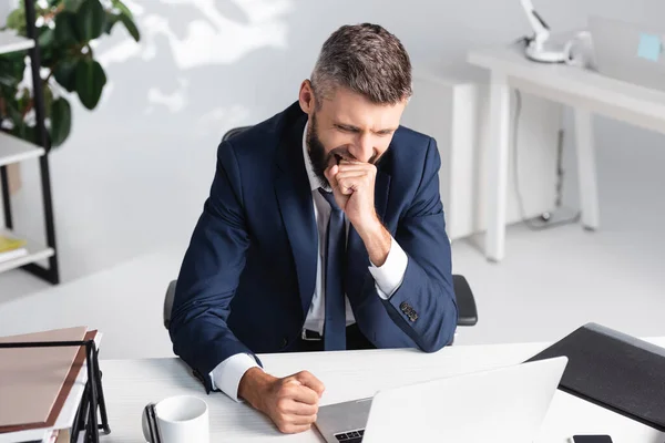 Empresario bostezando mientras trabajaba cerca de la computadora portátil y papeles en la mesa - foto de stock