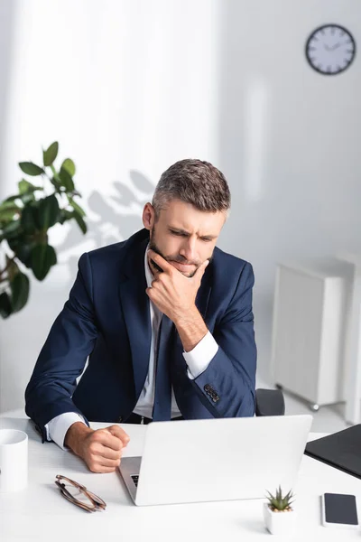 Pensativo hombre de negocios sentado cerca de gadgets y anteojos en primer plano borrosa en la oficina - foto de stock