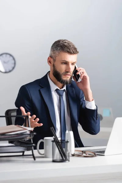 Hombre de negocios hablando en el teléfono inteligente cerca de la computadora portátil, taza y papelería en primer plano borrosa - foto de stock