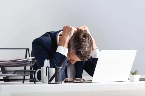 Hombre de negocios cansado sentado cerca de la computadora portátil y documentos en primer plano borroso en la oficina - foto de stock