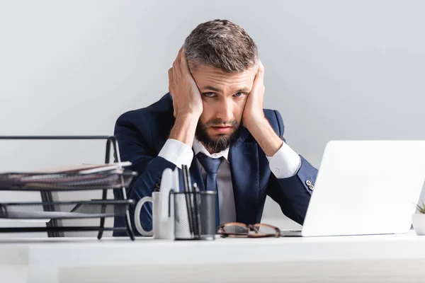 Uomo d'affari stanco guardando la fotocamera vicino laptop e cancelleria in primo piano sfocato — Foto stock