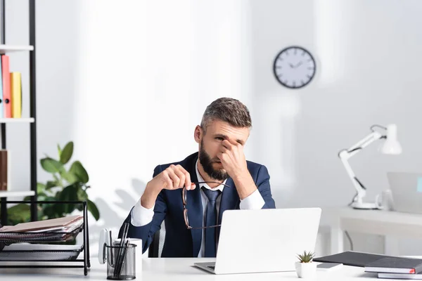 Businessman with hand near eyes holding eyeglasses beside laptop and stationery on blurred foreground on table — Stock Photo