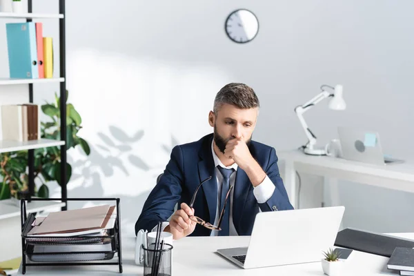 Empresario sosteniendo anteojos y mirando el portátil cerca de papeles y cuadernos en la mesa - foto de stock