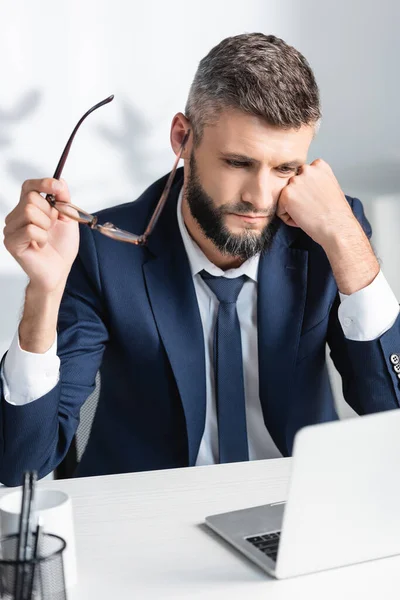 Empresário segurando óculos enquanto trabalhava com laptop em primeiro plano desfocado no escritório — Fotografia de Stock