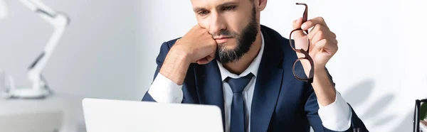 Empresario barbudo sosteniendo anteojos mientras mira el portátil en primer plano borroso en la oficina, pancarta - foto de stock