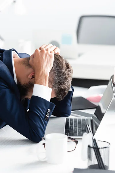 Overworked businessman touching head near laptop and stationery on blurred foreground — Stock Photo