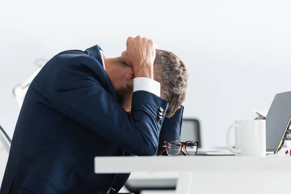 Empresário exausto de terno tocando a cabeça enquanto trabalhava perto de laptop, óculos e xícara no escritório — Fotografia de Stock
