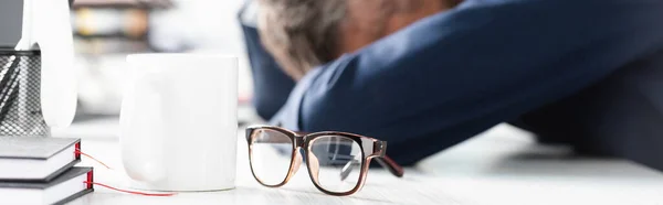 Очки и чашки на столе рядом с уставшим бизнесменом на размытом фоне в офисе, баннер — стоковое фото