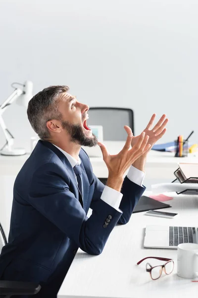 Angry businessman screaming near laptop and eyeglasses on blurred foreground on table — Stock Photo