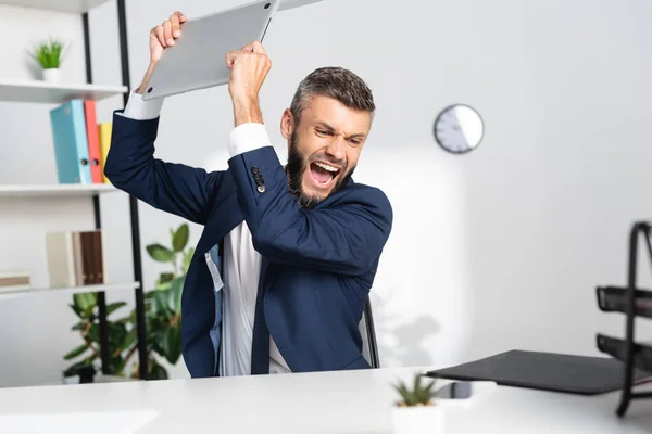 Uomo d'affari nervoso che tiene il computer portatile vicino a articoli di cancelleria in primo piano sfocati in ufficio — Foto stock