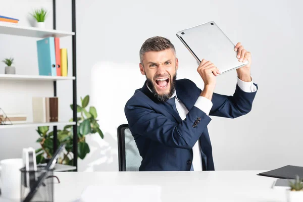 Irradiated and screaming businessman holding laptop near stationery on blurred foreground in office — Stock Photo