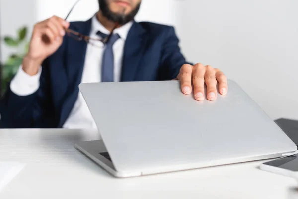 Vue recadrée de l'homme d'affaires touchant ordinateur portable tout en tenant des lunettes sur fond flou — Photo de stock