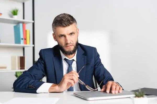 Empresario sosteniendo anteojos y mirando a la cámara cerca de la computadora portátil en primer plano borroso en la oficina - foto de stock