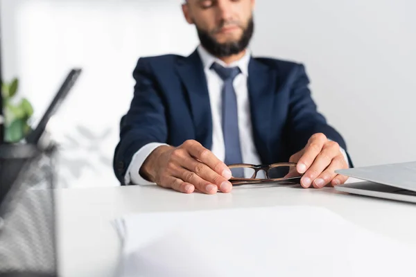 Vista ritagliata dell'uomo d'affari che tiene gli occhiali vicino; laptop in primo piano sfocato — Foto stock