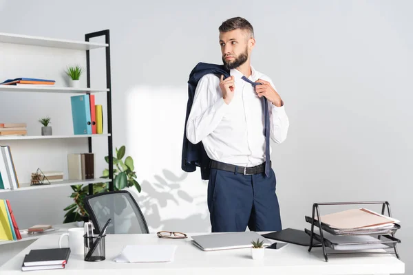 Empresario tocando corbata y sosteniendo chaqueta cerca de la computadora portátil y documentos en la mesa en la oficina - foto de stock