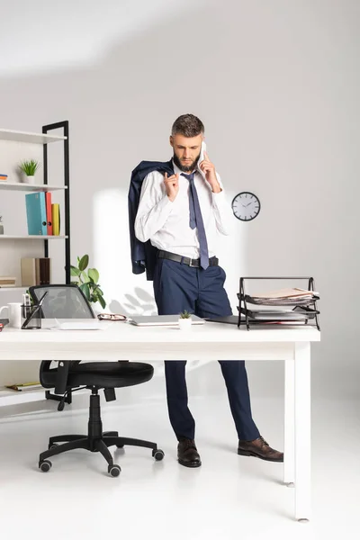 Empresario hablando en smartphone y sosteniendo la chaqueta cerca de los papeles en la mesa en la oficina - foto de stock