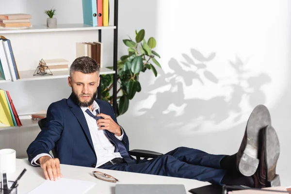 Businessman touching tie while looking at papers near laptop on blurred foreground in office — Stock Photo