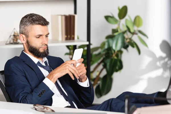 Businessman in suit using smartphone near eyeglasses on blurred foreground in office — Stock Photo