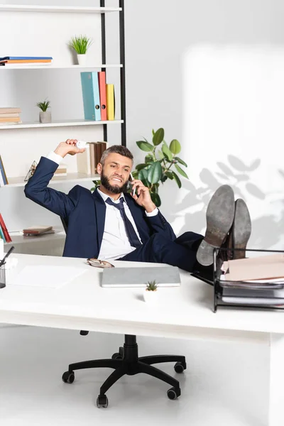 Empresario enojado sosteniendo papel agrupado mientras habla en el teléfono inteligente en la oficina - foto de stock