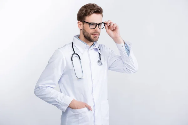 Уверенный в себе врач с рукой в кармане, держащий очки рамку, глядя в сторону изолированы на белом — стоковое фото