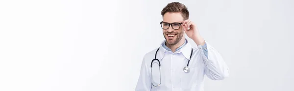 Feliz médico con estetoscopio, sosteniendo el marco de las gafas, mientras mira hacia otro lado aislado en blanco, bandera - foto de stock