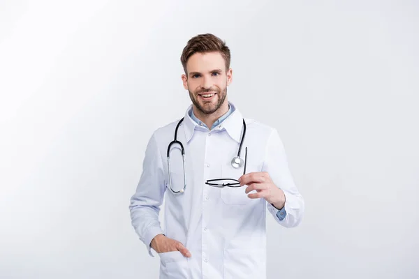 Vorderansicht eines lächelnden Augenarztes mit einer Hand in der Tasche, die eine Brille hält, während er die Kamera isoliert auf Weiß betrachtet — Stockfoto