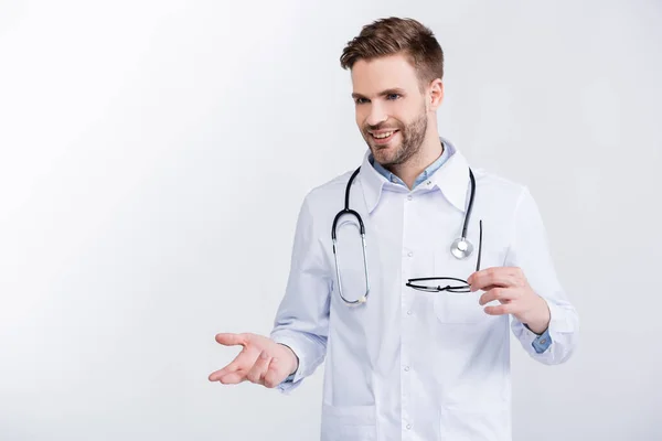 Sonriente médico adulto joven sosteniendo anteojos, haciendo gestos, mientras mira hacia otro lado aislado en blanco — Stock Photo