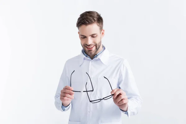 Médico sonriente mirando y sujetando un par de gafas con monturas negras aisladas en blanco - foto de stock