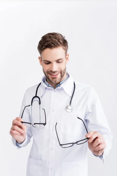 Médico sorridente olhando e segurando um par de óculos com molduras pretas isoladas em branco — Fotografia de Stock