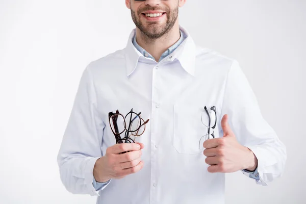 Vista recortada de médico sorridente com o polegar para cima, segurando vários óculos isolados em branco — Fotografia de Stock
