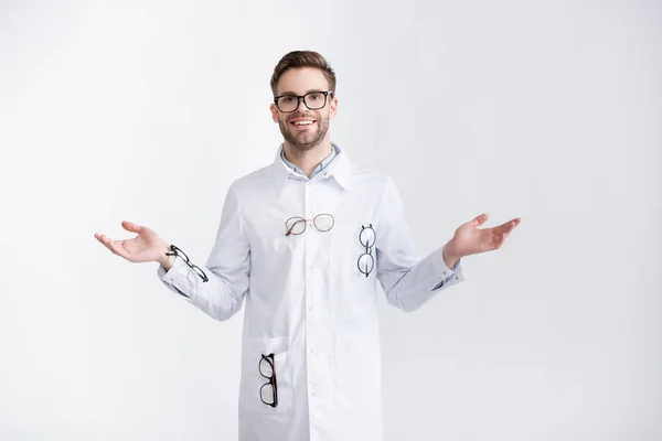 Vista frontal del oftalmólogo sonriente con los brazos abiertos con abrigo blanco con pares de anteojos colgantes aislados en blanco - foto de stock