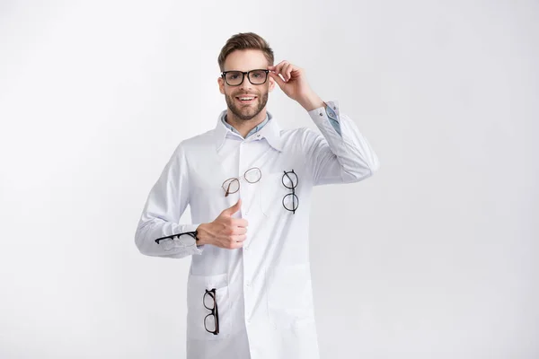 Vista frontal de oftalmólogo sonriente con el pulgar hacia arriba, con abrigo blanco con pares de anteojos colgantes aislados en blanco - foto de stock